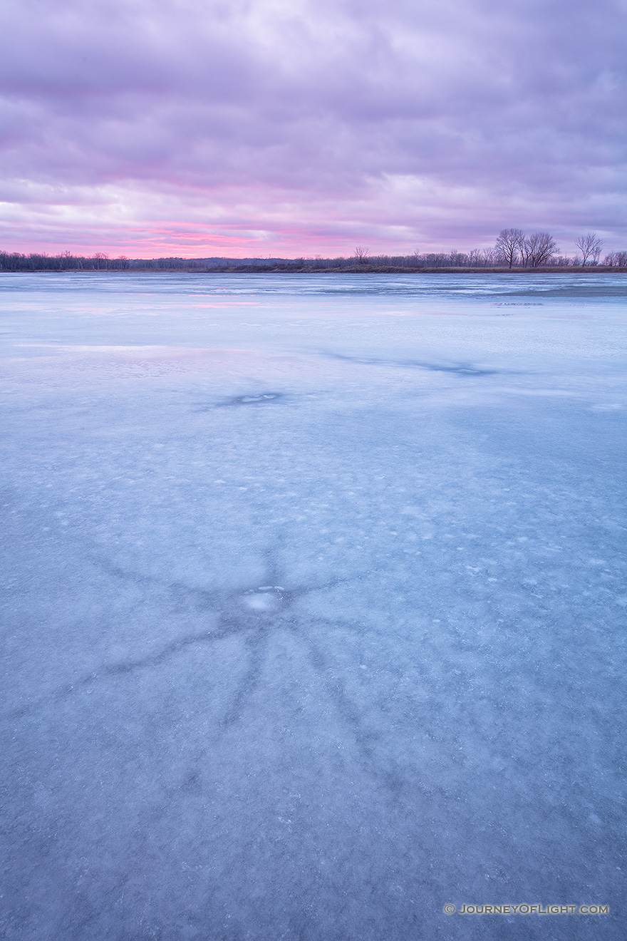 On this evening at DeSoto National Wildlife Refuge a quiet stillness prevailed.  I wandered the shore of the lake looking for interesting patterns in the now melting ice.  I found some interesting star shapes that had emerged from the recent thaws.  Sunset was near and although it was cloudy, I was hoping for a little illumination of the clouds.  After a while I gave up and begin leaving when a bit of sun broke free and I quickly set up again and captured this image. - DeSoto Picture
